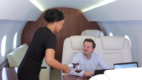 Work on laptop on board of private jet. Biracial flight attendant offering and pouring glass of wine for caucasian businessman and businesswoman travel inside of business airplane cabin.