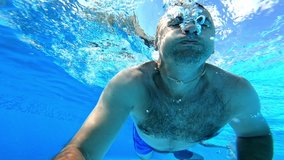 Swimmer swims forward while looking at camera underwater. Man swimming in clear water with underwater camera inside the swimming pool. Summer vacation.