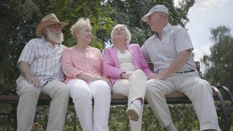 Two cute mature couples talking and smiling sitting on the bench in the summer park. Double date of senior couples. Friendly company resting outdoors. Old men and women met together. Retired people.