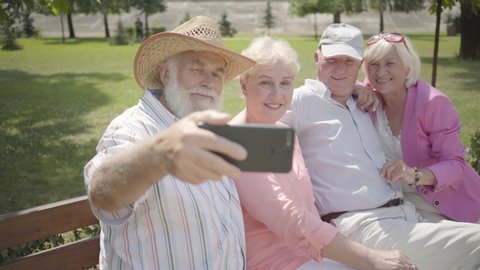 Two cute fun adult couples sitting on the bench together. The old man taking a selfie of company in the park. Mature people resting outdoors. Summertime leisure. Cheerful senior retired people.