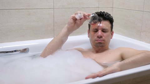 Adult drunk man lies in foam bath, spill red wine on his face a wine glass at home in the evening.