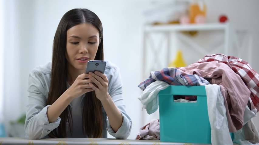 Smiling housewife chatting on smartphone near laundry basket with clothes