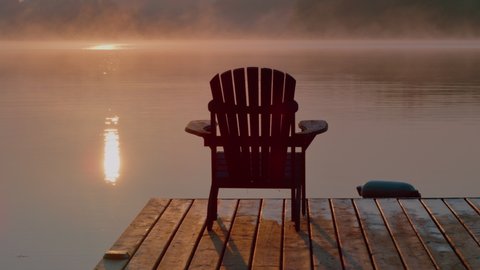 Muskoka chair on a dock at the cottage overlooking sunrise or sunset on the lake. Reflections shimmering in the water. Calm relaxing vacation.