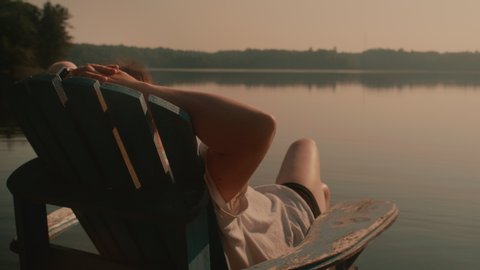 A woman sitting in a Muskoka chair on a dock, looking out on a lake. She is relaxing in the sunshine. The lake is calm and there are trees in the distance. 