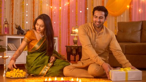 Diwali celebration - Handsome husband giving surprise gift to beautiful wife (Happy Indian Family). Indian Stock Footage of young couple wearing traditonal festive dress sitting on floor near rango...