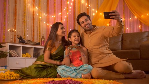 Diwali festival - Indian lovable family taking selfie or self photograph at home. Colorful decorate background. Asian/Indian young family taking photographs at an apartment with a plate of rose pet...