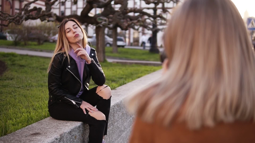 Footage from the shoulder - professional female photographer takes pictures of a girl model her friend. Attractive long haired woman in black casual clothes posing for the camera, sitting on the