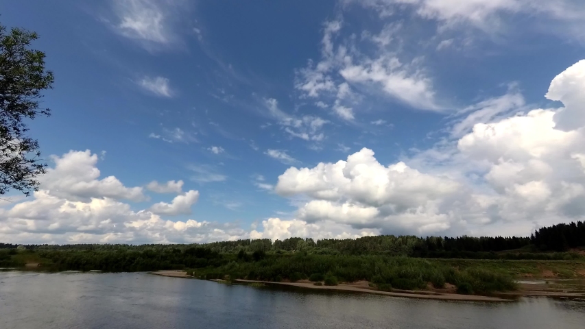 Clouds run across the blue sky. Time lapse. Below the river flows and the forest grows
