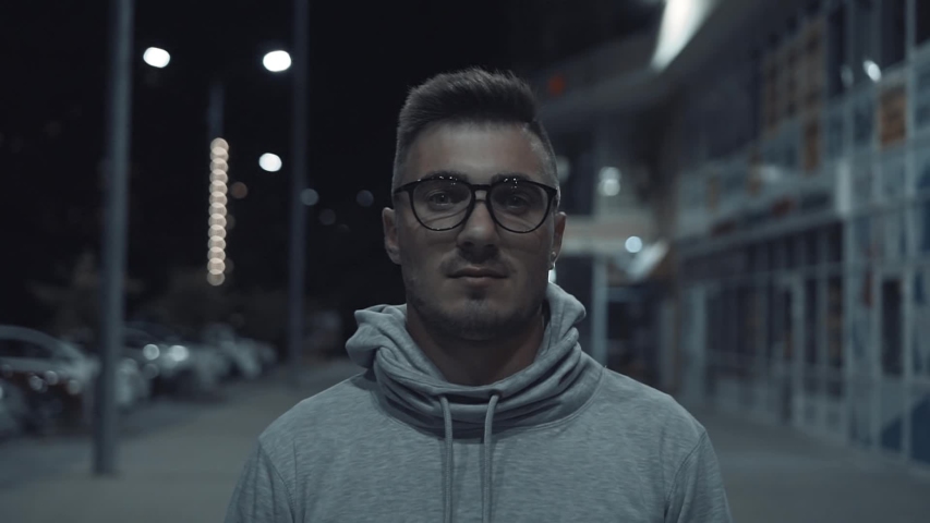 A happy and handsome guy with glasses looks into the frame and smiles. It is located on the night street of the city.