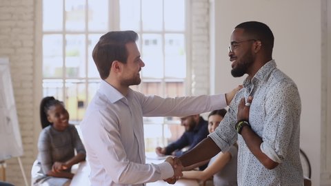 Proud caucasian manager boss handshaking congratulating praising male black african worker give positive feedback supporting promising reward promoting at team meeting, employee recognition concept