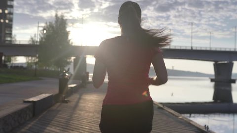 Person running in city. Woman on a morning jog. Following slow motion back view. Fitness runner training. Sunny sky with clouds at sunset or sunrise. Jogger on dock by the lake. Sport motivation.