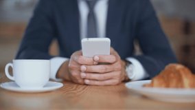 cropped view of businessman using smartphone while sitting in cafe near cup of coffee and plate with croissant