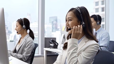 Young asian woman wearing microphone headset working in customer service support help desk with colleagues in call center office