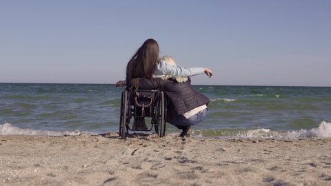 Young disabled woman in wheelchair with her mother hugs each other on the sand beach near the sea