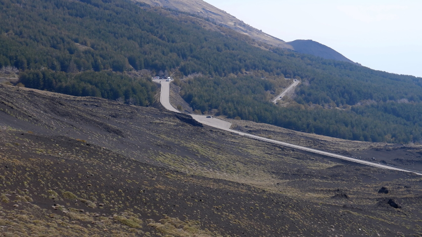 Panoramic view of cars driving winding SP92 road towards mountain refuge, the Sapienza Refuge, Etna volcano base and people at the top of extinct Silvestri Superiore crater.