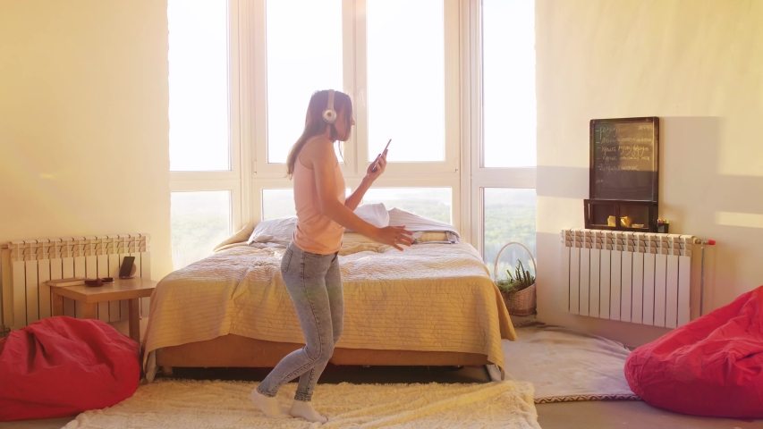 Happy brunette young woman in headphones dancing alone in bedroom with big window listens to music on smartphone, girl holding phone and falls on the bed having fun at home. slow motion.
