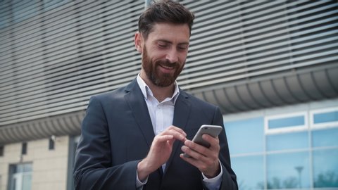CloseUp of Attractive Young Businessman using Smartphone and Smiles. Texting Messages while standing near Modern Office Building. Wearing Classical Suit. Social Network. Apps. Smartphone.