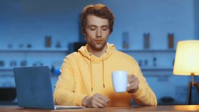front view of blogger with laptop drinking tea and talking to camera in kitchen