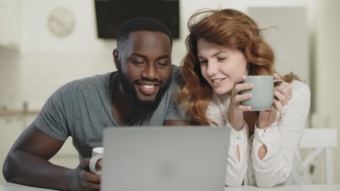 Black man talking woman at home. Happy couple having conversation at table near laptop. Smiling man and woman reading news at home in morning