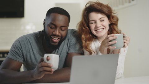 Happy couple talking at open kitchen with tea. Diverse family looking laptop at open kitchen. Black man and white woman sitting on table at living room.