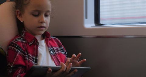 Cute Little Girl Entertaining With Tablet. Child Spending Leisure Time, Rides an Electric Train, Playing Mobile Game in the and Crushes the Bright Screen With Her Hand. Happy Family.