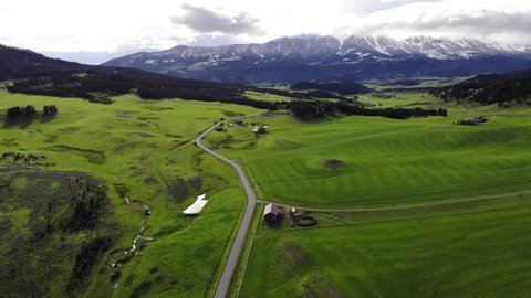 Drone shot over a road and green fields with snowy mountains ahead.