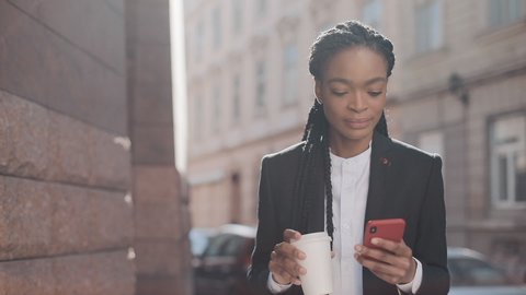 Portrait of a young African American businesswoman in a suit, walking around the city, drinking coffee and using smartphone. Female professional in suit having good news on smartphone.