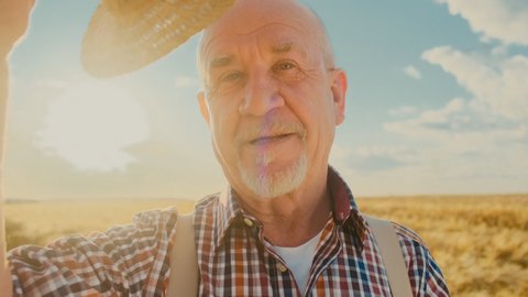 Close up of the old Caucasian farmer man in a plaid shirt doing a greeting movement or gesture with a hat and looking straight to the camera in the field. Portrait.