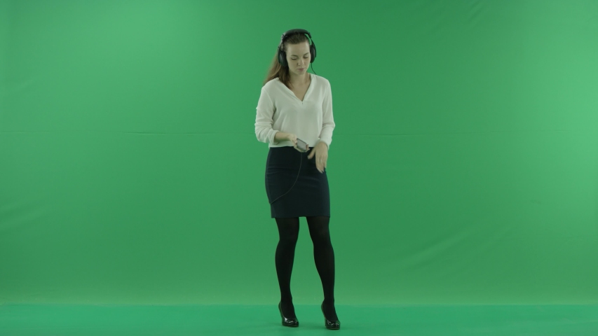 Happy businesswoman listening to music in the headphones and dancing on the spot. She wears formal dress: white shirt and blak skirt, standing in the centre the greenscreen