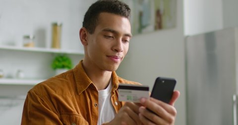 Young latin man is doing online shopping, using credit card and smartphone, cheerful and smiling, being in raised mood at home, close up. 4K, shot on RED camera.