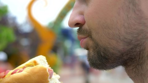 young man eating an appetizing hotdog sitting in the park on the background of walking people, blurred background.