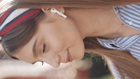 Vertical video. Portrait of a beautiful young woman with wireless earphones looking into the camera and smiling standing on the old street background. Girl wearing in striped shirt dress.