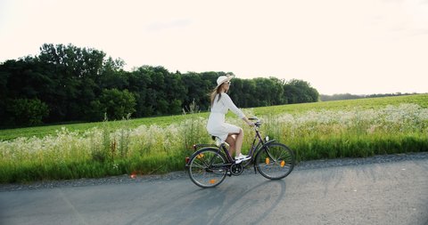 Glamorous lady in white dress, hat and sunglasses riding bicycle in countryside and having fun, feeling happy and inspired