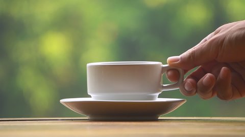 Close-up of hand placing hot ceramic white coffee cup with smoke on saucer over wooden table in nature green background