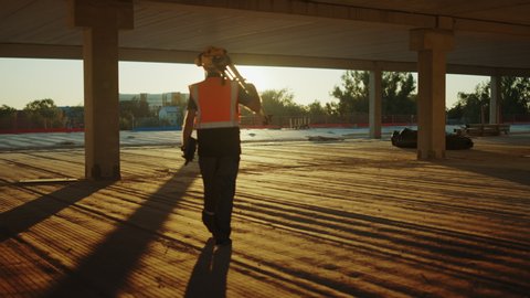 Inside of the Commercial / Industrial Building Construction Site: Professional Engineer Surveyor Finishes Day's Job Carries Tripod Theodolite and Walks into the Sunset