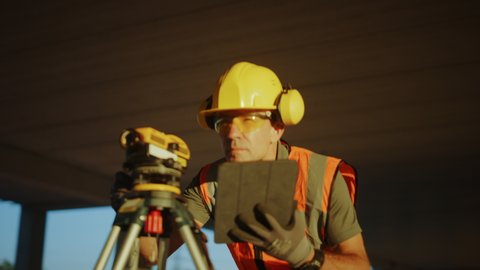 Inside of the Commercial / Industrial Building Construction Site: Professional Engineer Surveyor Takes Measures with Theodolite, Using Digital Tablet Computer