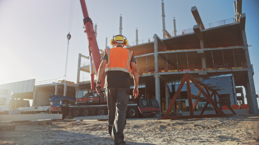 Following Shot: Worker Contractor Wearing Hard Hat and Safety Vests Walks on Industrial Building Construction Site. In the Background Crane, Skyscraper Concrete Formwork Frames Royalty-Free Stock Footage #1034288204