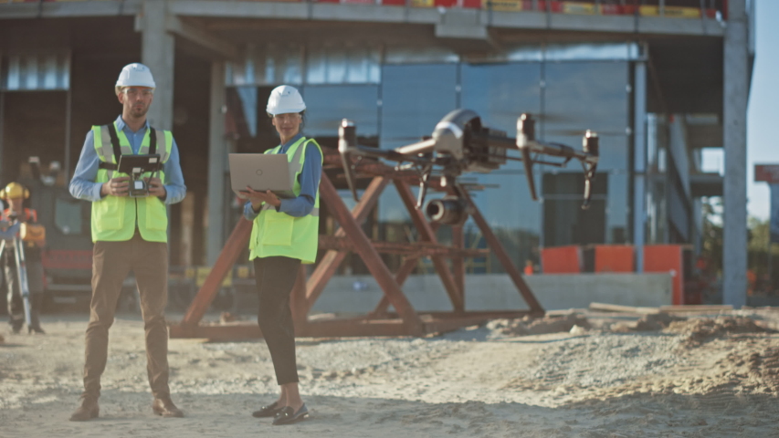 Diverse Team of Specialists Pilot Drone on Construction Site. Architectural Engineer and Safety Engineering Inspector Fly Drone on Commercial Building Construction Site Controlling Design and Quality Royalty-Free Stock Footage #1034288246
