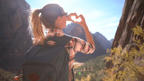 Young woman on top of mountain making heart shape finger frame with hands loving nature and embracing environment; Hiking girl enjoying outdoor activities 