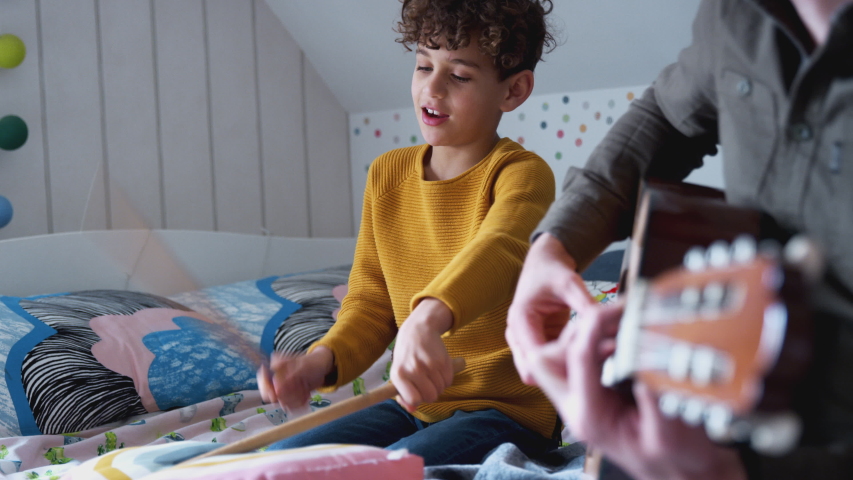 Single Father Playing Guitar With Son Who Drums On Cushion In Bedroom