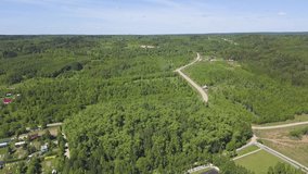 Aerial view of suburban area with many green trees and a road under the summer sky. Clip. Small town among forested hills on blue sky background.