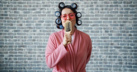 Woman with hair curlers, glasses and bathrobe is singing in hairbrush having fun moving arms standing on brick wall background. People, joy and music concept.