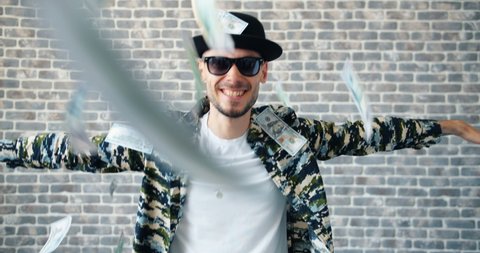 Slow motion of young hipster businessman dancing on brick wall background when money is falling and flying around. Happy people, finance and success concept.