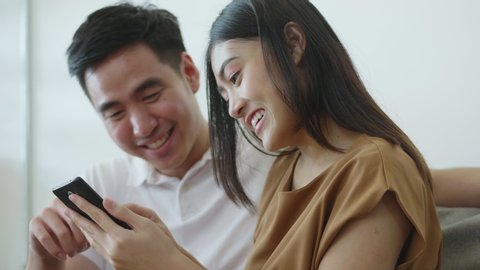 Slow Motion: Close up Asian couple using smartphone browsing social media communication and shopping online while sitting on the sofa at home.