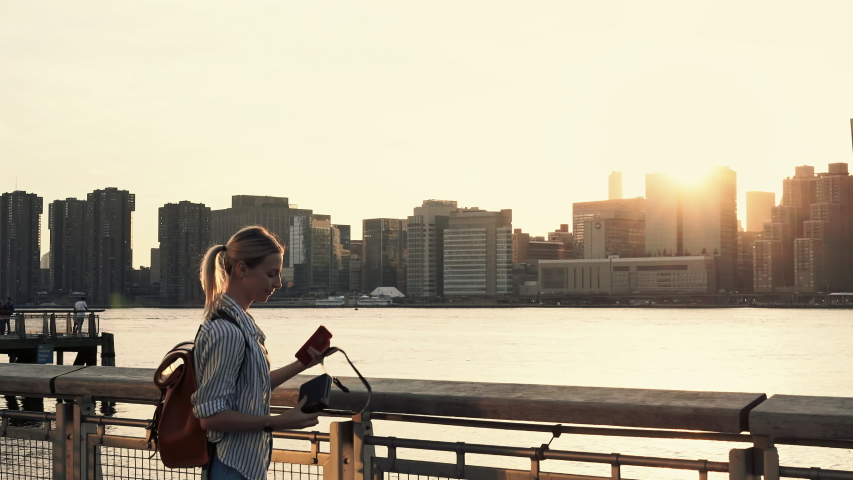 Hipster girl photographing scenic landmark of Manhattan island via film instant camera looking at made image and clicking photo with smartphone application for sharing to travel website
