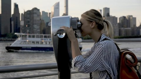 Attractive female traveller with touristic backpack using Tower Optical viewer for looking around metropolitan skyline buildings, young hipster girl with retro technology on neck exploring city