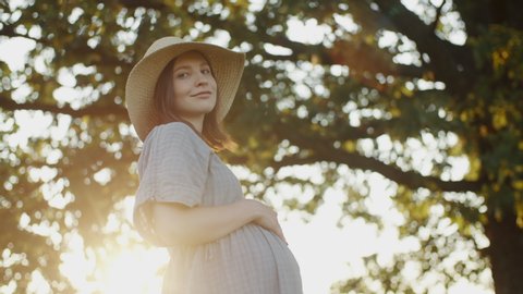 Happy pregnant woman touches her belly dressed in linen dress and straw hat standing by oak tree in meadow during sunset. Beautiful expectant mother looks into camera smiles in evening sunlight flares
