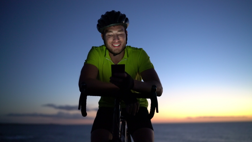 Cyclist biking looking at mobile phone while cycling riding road bike. Athlete biker using smartphone app on workout at sunset. Light from screen on face.