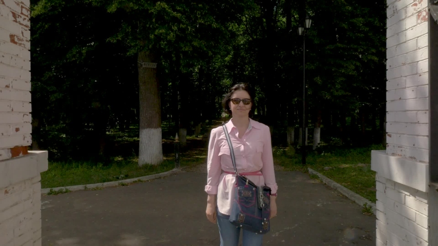 A middle-aged woman, a brunette in a pink shirt with a red belt and a bag over her shoulder, go through the gate from an old park on a summer day. medium shot. Camera moving