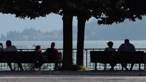 Cernobbio, July, 2019 – group of elderly people rest and talk sitting on the benches by the lake with camera zoom out
